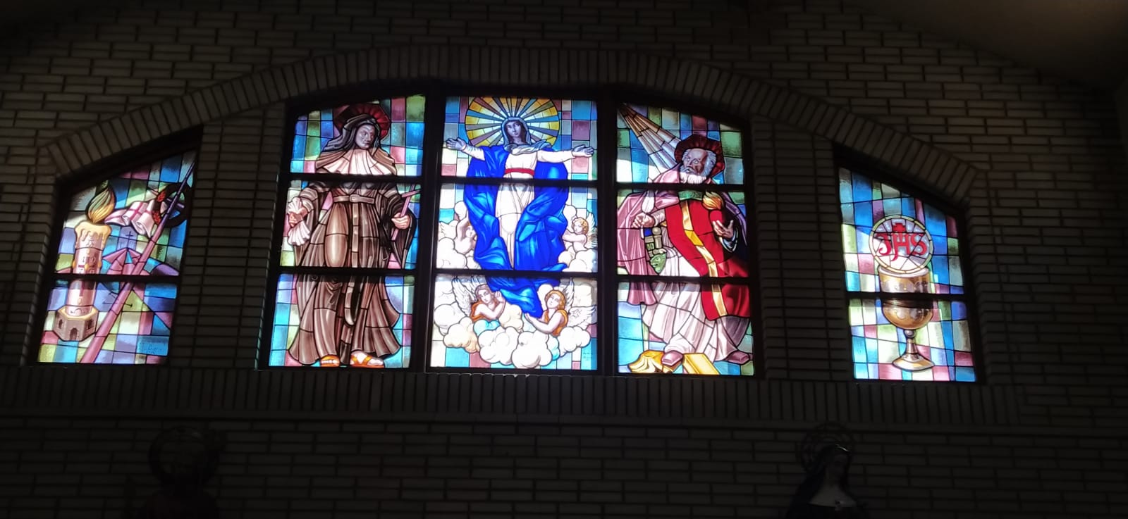En el centro de la vidriera, encontramos la Asunción de María. Junto a ella, aparece la imagen de Santa Mónica y su hijo San Agustín. A ambos lados, vemos las alegorías de la Eucaristía y el Cirio Pascual.